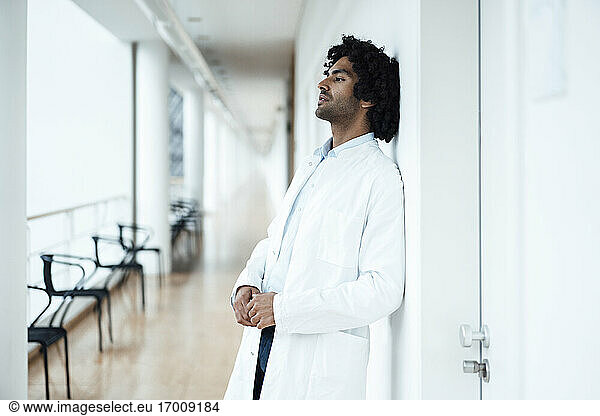 Nachdenklicher junger Mann im Gesundheitswesen lehnt an einer weißen Wand im Krankenhausflur