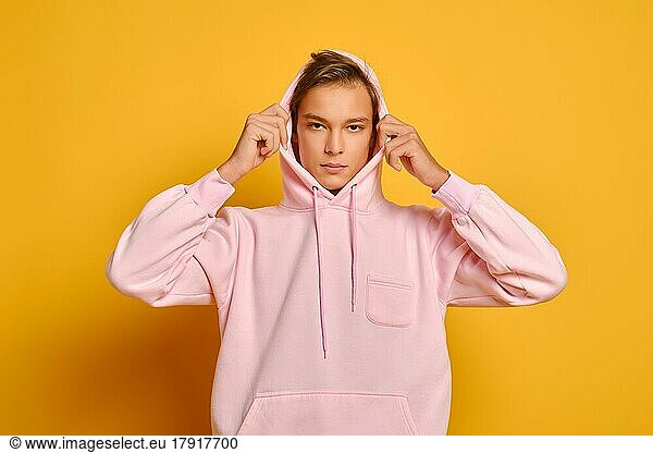 Nachdenklicher junger Mann in rosafarbenem Sweatshirt setzt Kapuze vor gelbem Studiohintergrund auf