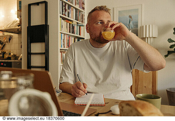 Nachdenklicher Mann trinkt Saft und schreibt zu Hause in sein Tagebuch