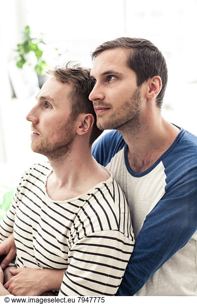 Nachdenkliches junges homosexuelles Paar schaut weg  während es sich zu Hause umarmt.