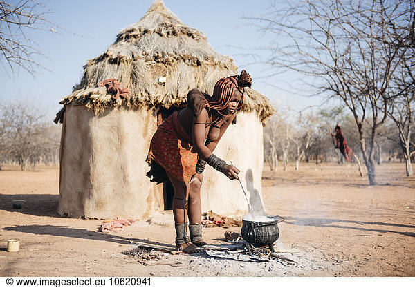 Namibia  Damaraland  young woman cooking on a small fire in front her hut in a Himba village