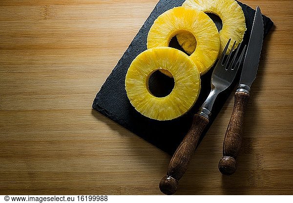 Natürliche Ananasscheiben auf einer Schieferplatte und etwas Besteck.
