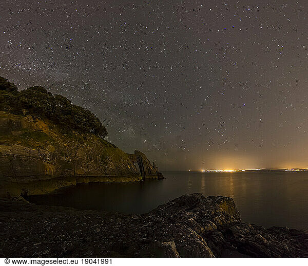 Natural sea arch and coastline at night with starry sky and lights