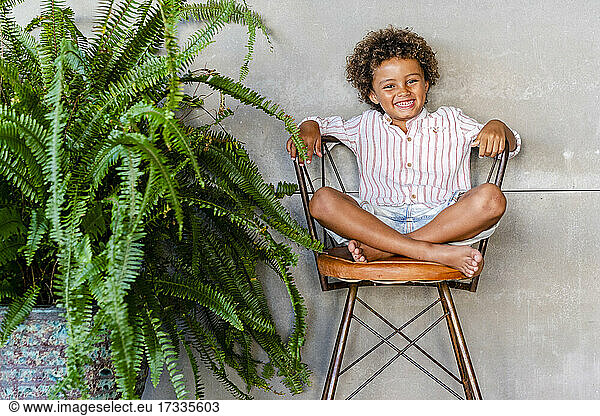 Netter lächelnder Junge sitzt auf einem Stuhl bei einer Pflanze zu Hause