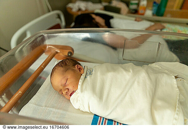Newborn baby in swaddle sleeping in hospital room near mother.
