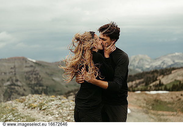 newly engaged white couple dressed in all black kiss on windy ridge