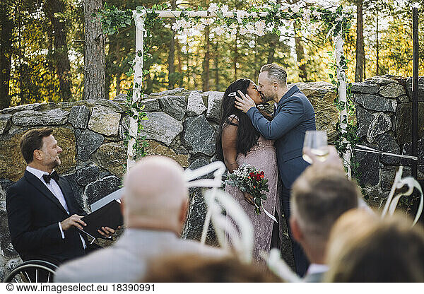 Newlywed couple kissing in front of guests and minister at wedding ceremony
