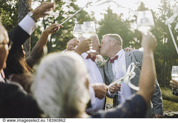 Newlywed gay couple kissing in front of guests raising toasts at wedding ceremony