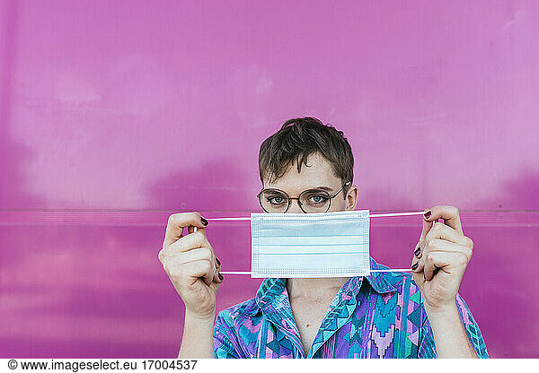 Nicht-binäre Person  die eine schützende Gesichtsmaske hält  während sie an einer rosa Wand steht