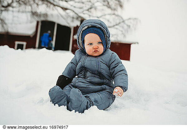 Niedliches Baby sitzt auf Schnee und berührt zum ersten Mal Schnee auf dem Bauernhof