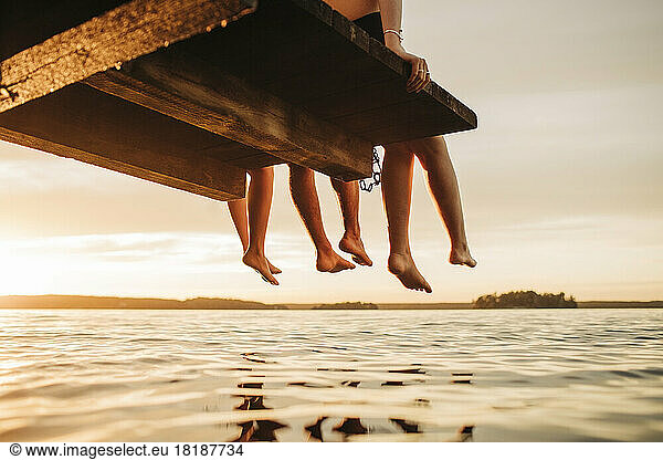 Niedriger Ausschnitt von männlichen und weiblichen Freunden  die bei Sonnenuntergang auf einem Steg am See sitzen