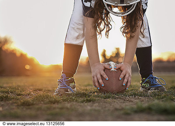 Niedriger Teil eines Mädchens  das American Football in der Hand hält  während es bei Sonnenuntergang auf einem Rasenfeld gegen den Himmel steht