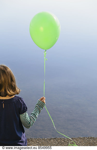 Nine year old girl holding green balloon, sitting by waters edge, Nine year old girl holding green balloon, sitting by waters edge