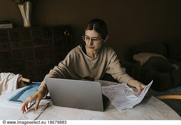 Non-binary person collecting bills while sitting with laptop at home