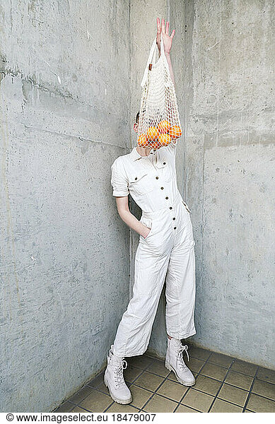 Non-binary person holding bag of oranges standing in front of wall