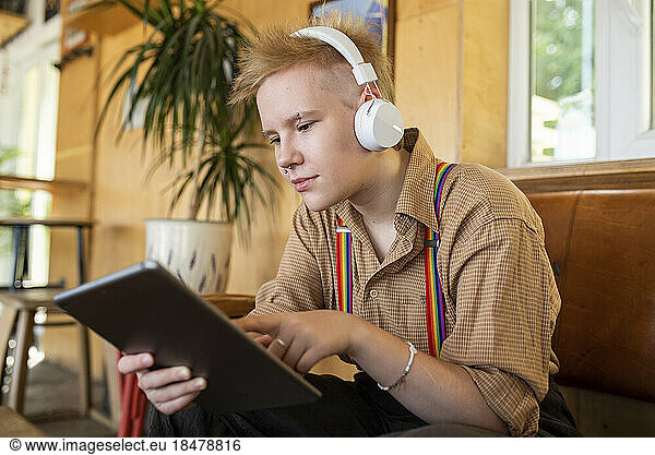 Non-binary person using tablet PC wearing headphones listening to music at cafe