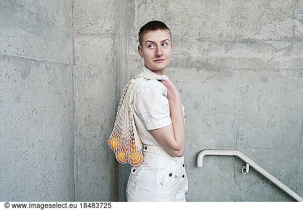 Non-binary person with bag of oranges standing by wall