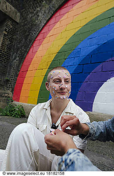 Non-binary person with sticker on face sitting by friend in front of multi colored wall