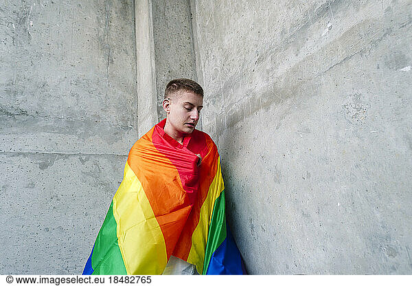 Non-binary person wrapped in rainbow flag by wall