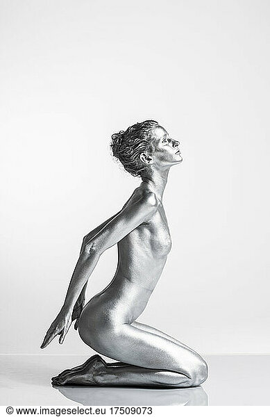 Nude Caucasian woman with silver body paint kneeling  hands behind her