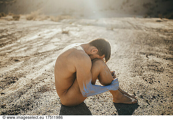 Nudist man with body paint hugging knees while sitting at desert during sunny day
