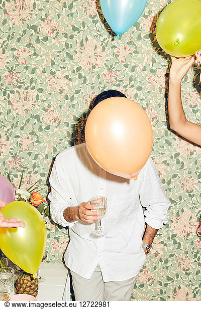 Obscured face of young man holding drink while standing by woman with balloons against wallpaper at home during dinner p