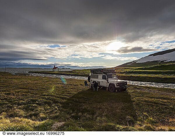 Off-road car parked in front of Icelandic river at cloudy sunset with secluded Unadsdalskirkja church in background