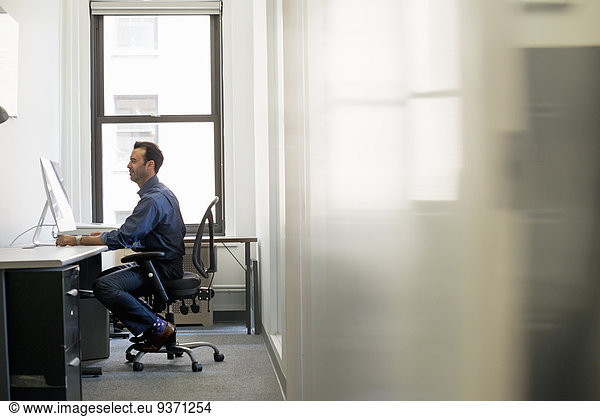 Office life. A man in casual clothing seated at a desk looking at a computer screen.