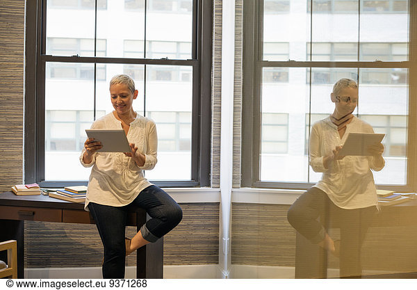 Office life. A woman seated on the edge of her desk using a digital tablet.