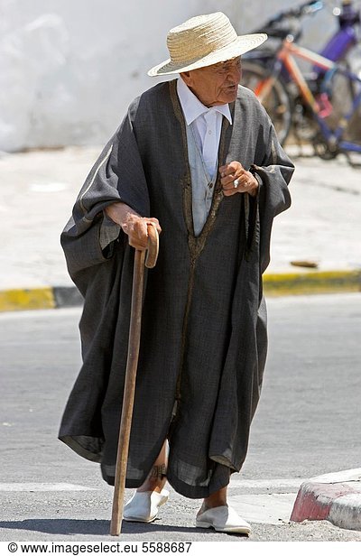 Old Berber man in traditional woollen cloak and straw hat walks with stick in street Matmata Tunisia
