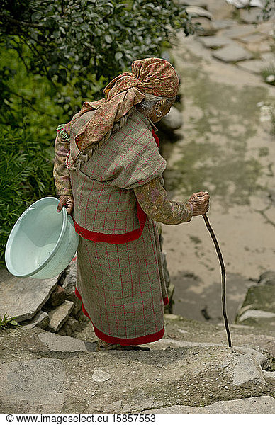 Old Kullu woman walks down a staircase with a cane and plastic basin