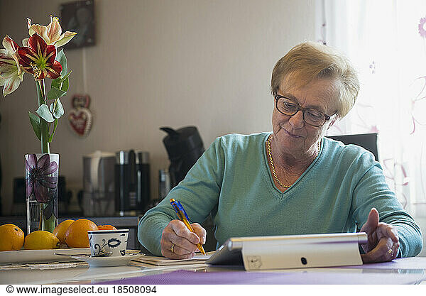 Old woman watching digital tablet and writing in book at dining table