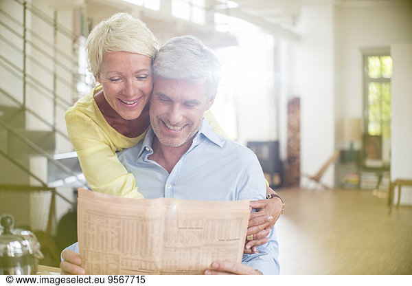 Older woman hugging husband reading newspaper