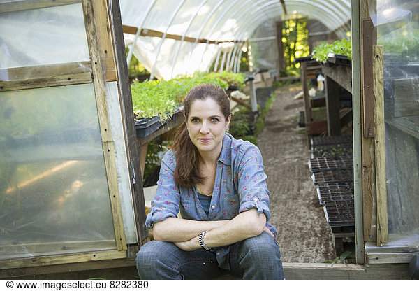 On The Farm. A Woman Sitting Resting At The Door Of A Glasshouse With Benches Full Of Young Seedling Plants.