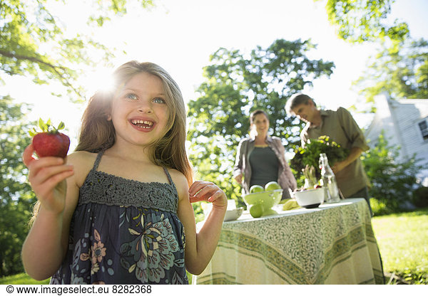 On The Farm. Children And Adults Together. A Young Girl Holding A Large Fresh Organically Produced Strawberry Fruit. Two Adults Beside A Round Table.