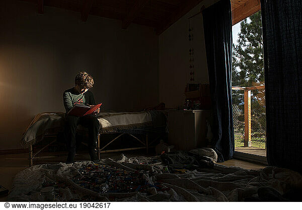 One boy sitting on his bed holding a notebook in a room with legos