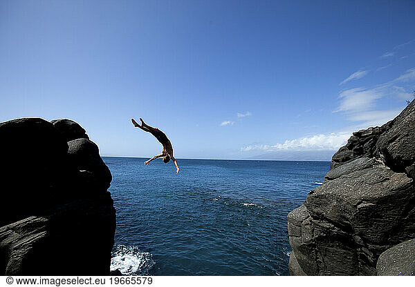 One man back flipping off of a cliff into the ocean on a blue sky day.