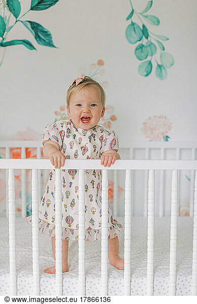 One year old girl in pink dress stands in crib and smiles