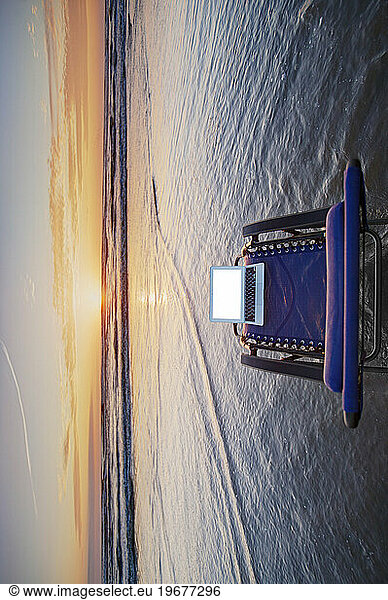 Open laptop on sun lounger in shallow wave at beach at sunset.