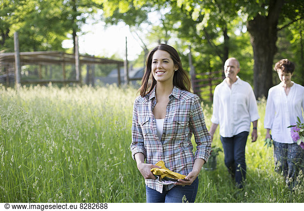 Organic Farm. Summer Party. Two Mature People  A Couple And A Young Woman Walking Through The Long Grass.