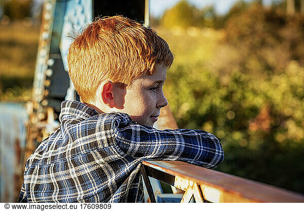 Outdoor portrait of a boy with red hair standing on a bridge in a park looking out over the railing; Edmonton  Alberta  Canada