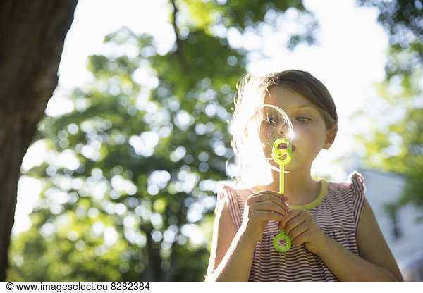 Outdoors In Summer. A Young Girl Blowing Bubbles In The Air Under The Branches Of A Large Tree.
