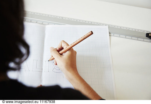 Over the shoulder view of a woman working on a graphic on a drawing board  outlining letters with a pencil.
