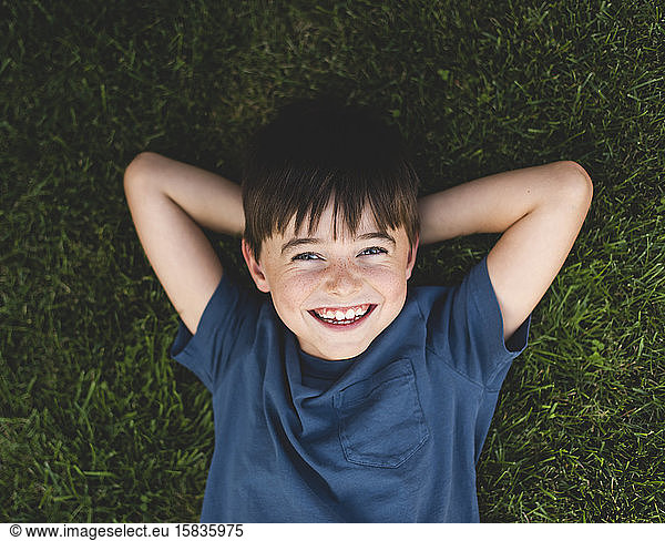 Overhead close up of happy boy laying on grass with arms behind head.