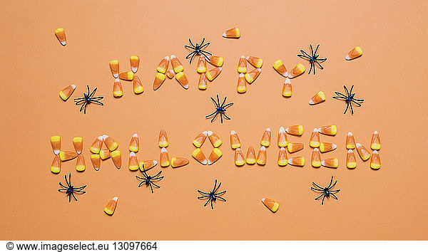 Overhead view of candy corns arranged as Happy Halloween text amidst artificial spiders over orange background