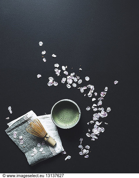 Overhead view of matcha tea and bamboo whisk by petals on black background