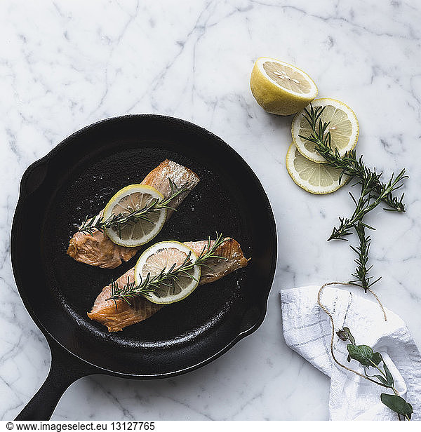 Overhead view of salmon served in cooking pan by lemon and rosemary on table