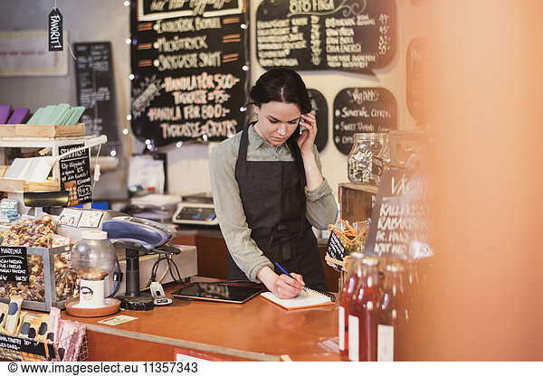 Owner talking on phone while writing on note pad at checkout counter in store