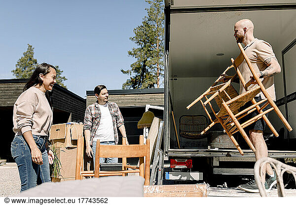 Pärchen sieht zu  wie ein Möbelpacker an einem sonnigen Tag Stühle vom Lastwagen ablädt
