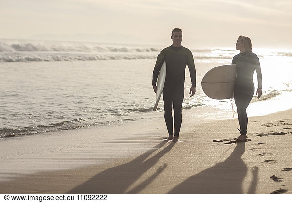 Paar mit Surfbrettern  die bei Sonnenuntergang am Strand spazieren gehen.